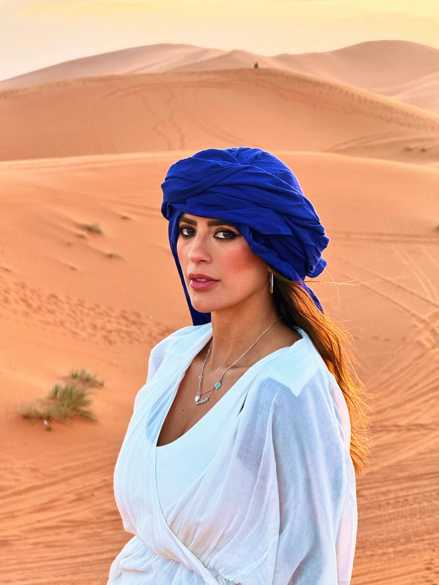 Woman with blue headscarf and white dress in desert with sand dunes, sunset sky.