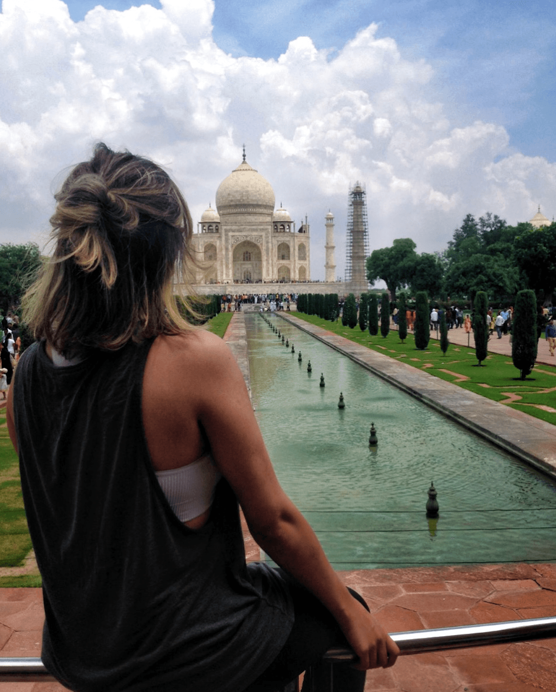 Person with shoulder-length hair looking at the Taj Mahal with a water feature in front, cloudy sky above.
