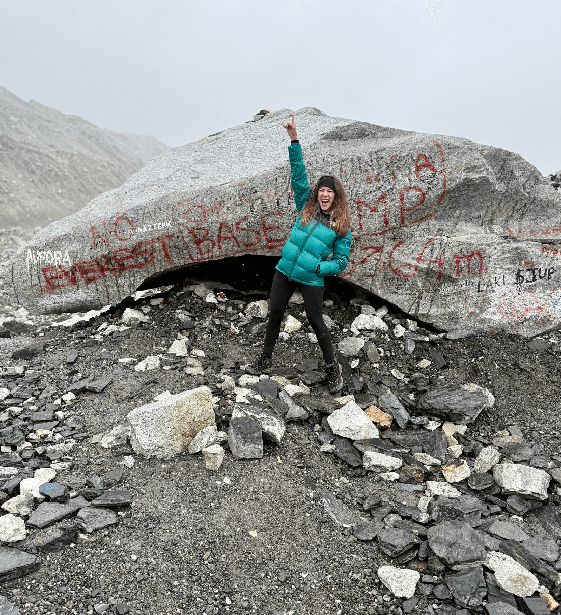 Person standing on rocky ground in front of a large boulder with graffiti, holding a stick or pole, wearing a teal jacket.