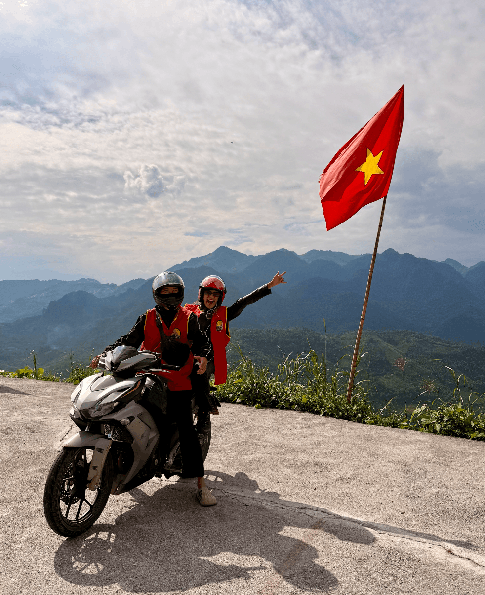 Two people wearing helmets and red vests stand next to a motorcycle on a mountain road, with a Vietnamese flag and mountain landscape in background.