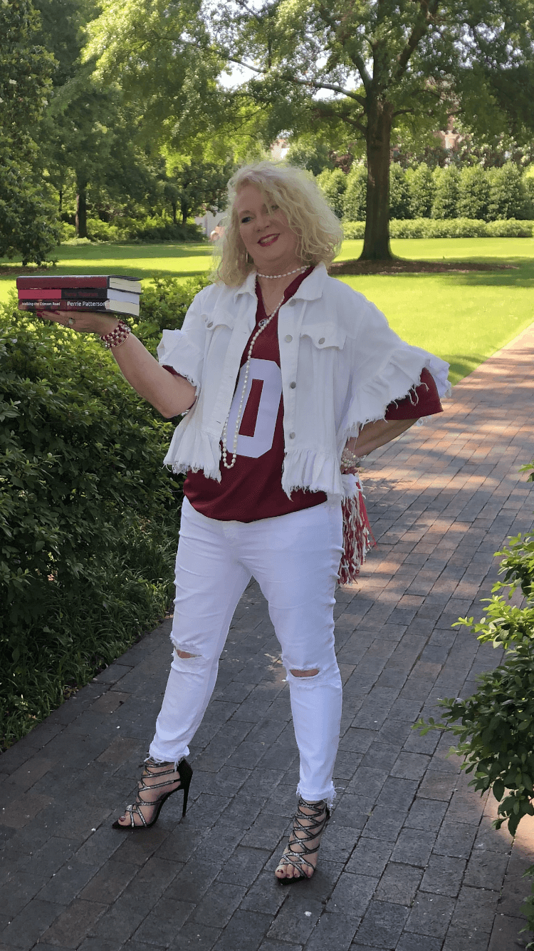 Woman with blonde curly hair standing outdoors on a paved path, holding a stack of books, surrounded by trees and greenery.