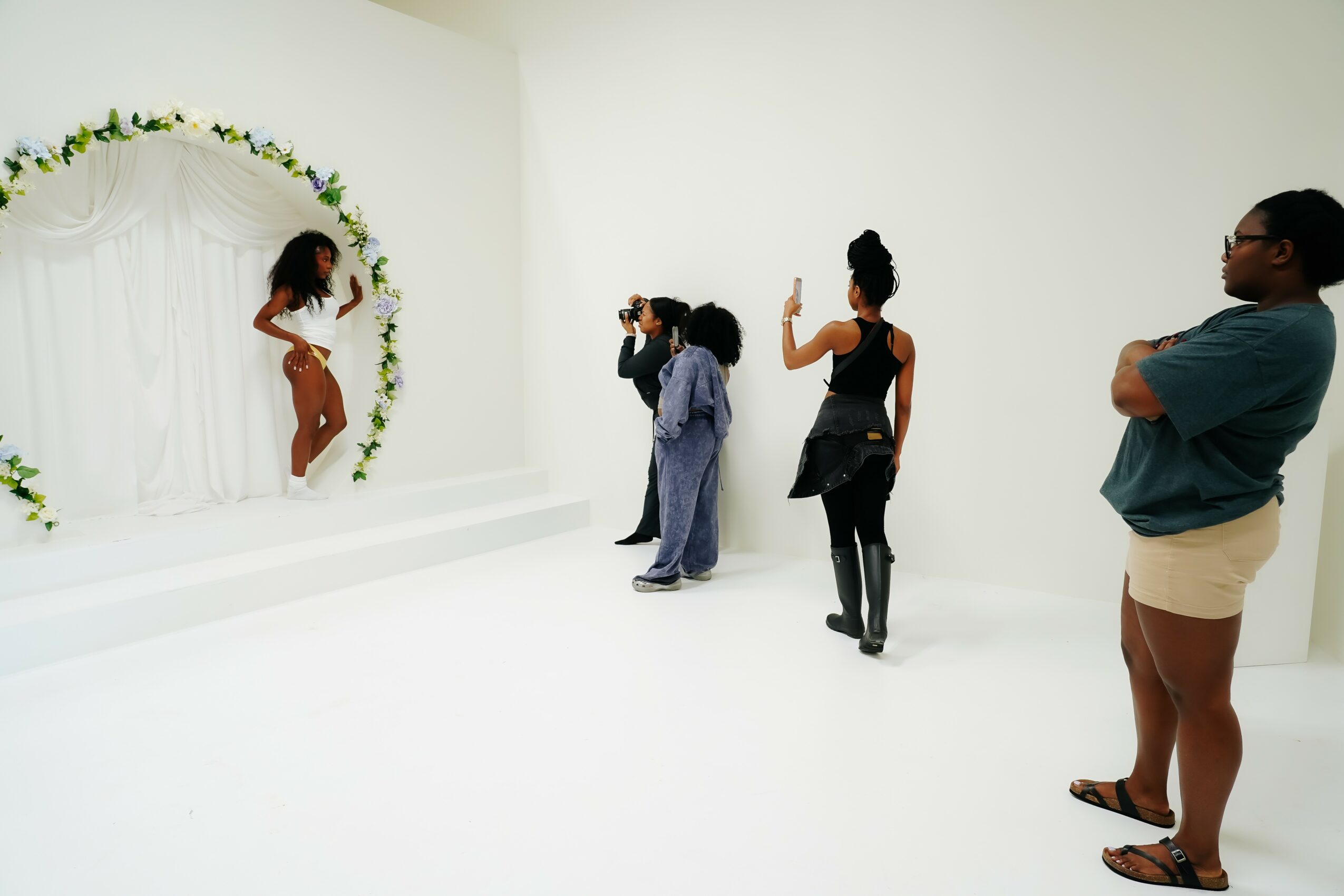Group of people taking photos of a woman in a floral arch, with a white background and minimal decor.