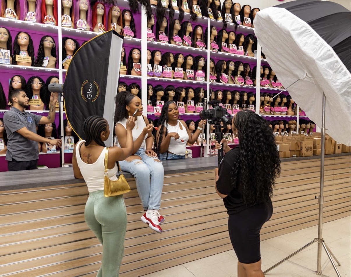 People in a wig store with mannequins and wigs on shelves, a photographer, and a woman being photographed, with studio lighting equipment.