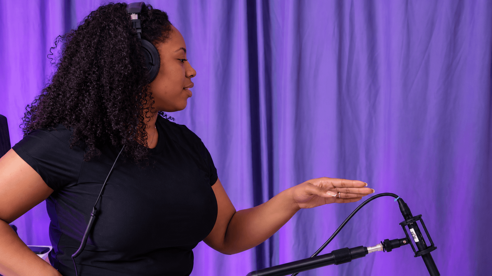 Woman with curly hair wearing headphones, gesturing with her right hand, purple curtain background.