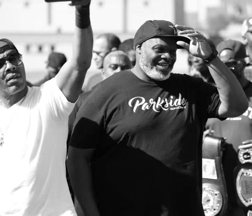 Two men smiling outdoors, one wearing a cap and a t-shirt with 'Parkside' written on it, in black and white.