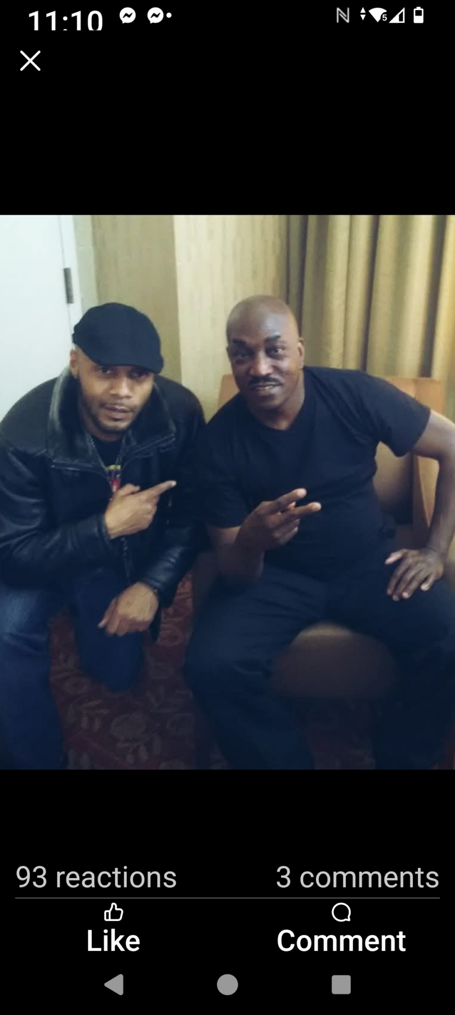 Two men sitting on a sofa, one wearing a black jacket and cap, the other in a black shirt, both making hand gestures.