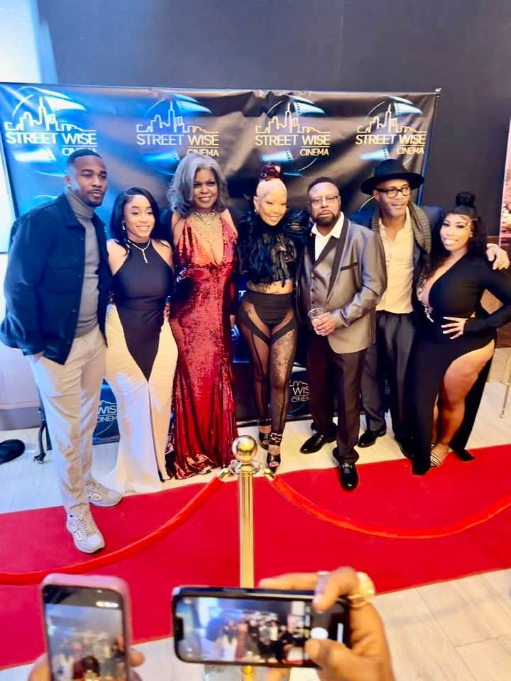 Group of seven people posing on red carpet in front of a black backdrop with logos, some holding phones, dressed in formal attire.