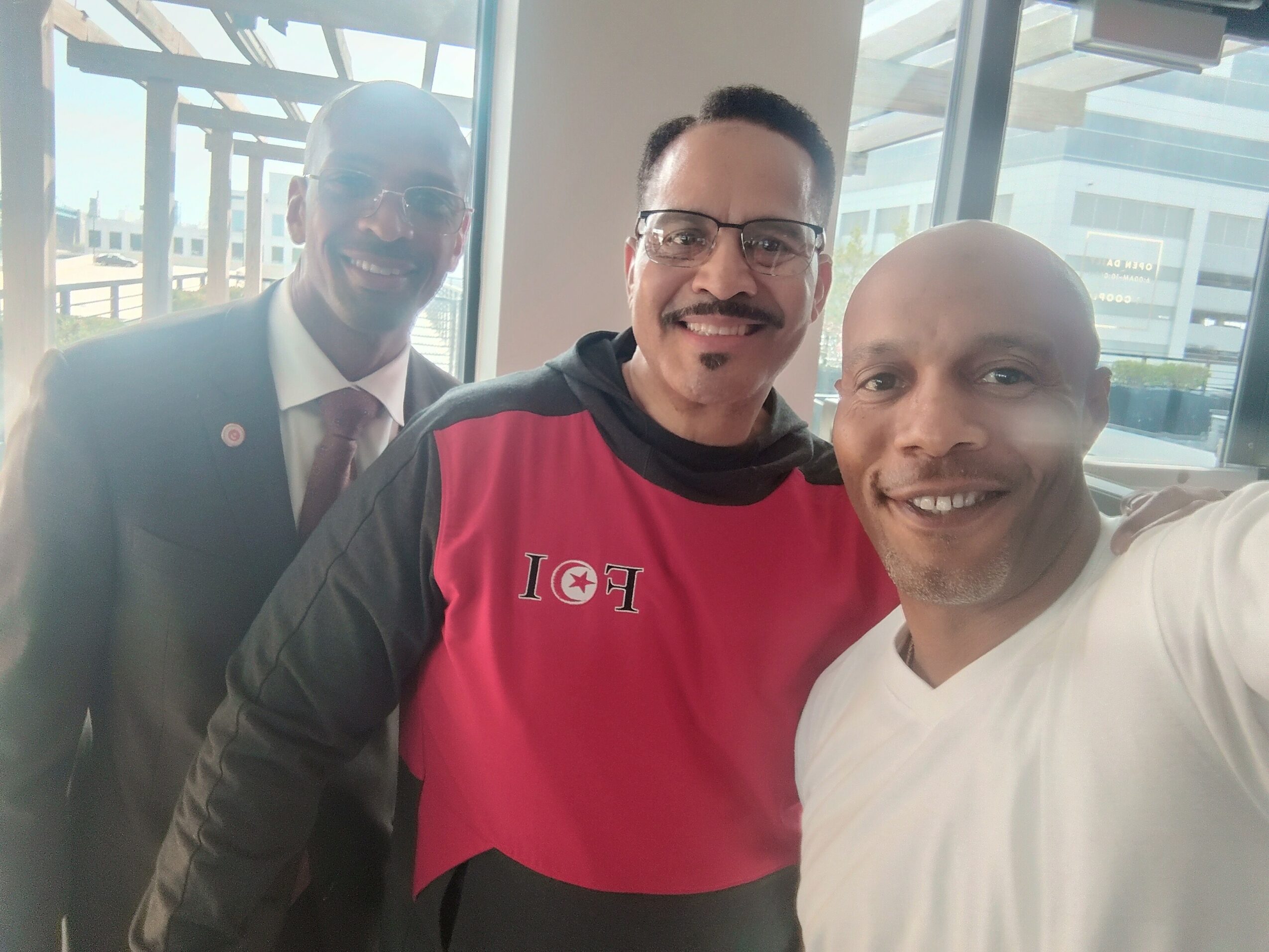Three men smiling indoors with large windows and buildings outside, one wearing a suit, one in a red and black shirt, and one in a white shirt.