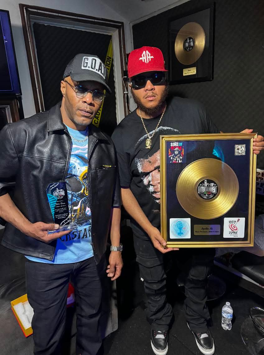 Two men in a recording studio, one holding a framed gold record plaque, both wearing sunglasses and caps.