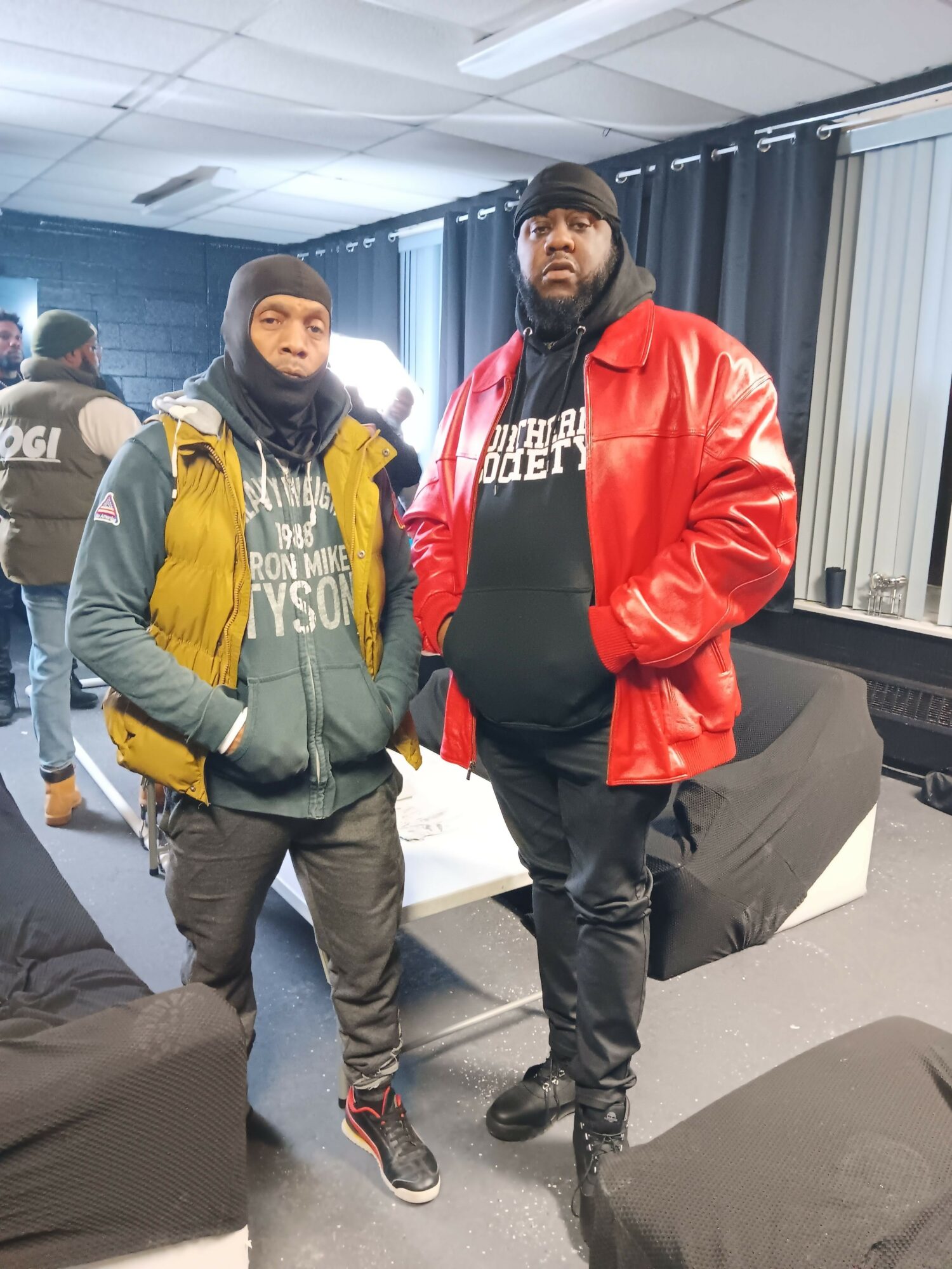 Two people standing indoors, one in a red jacket and black pants, the other in a yellow jacket and gray pants.