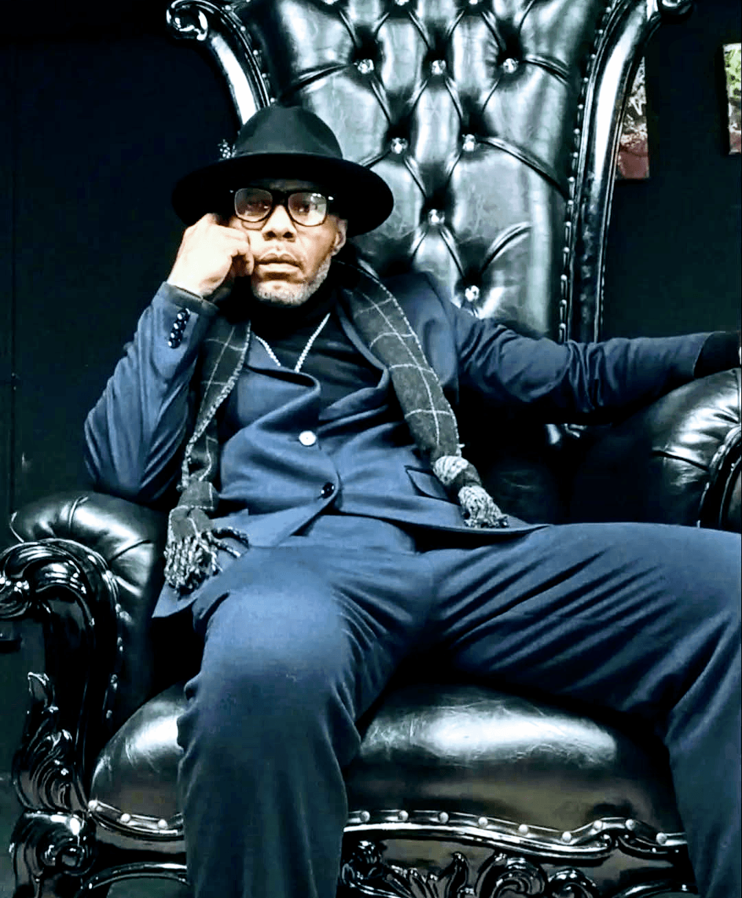 Person sitting on a large black leather chair, wearing a hat, glasses, and dark clothing, with a serious expression.