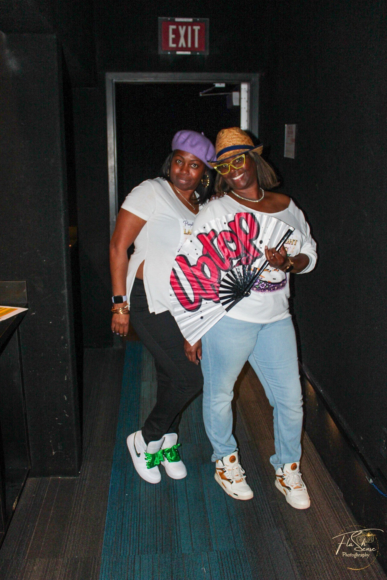 Two women stand together in a dark hallway, smiling, wearing casual clothes and colorful accessories, with an exit sign above them.