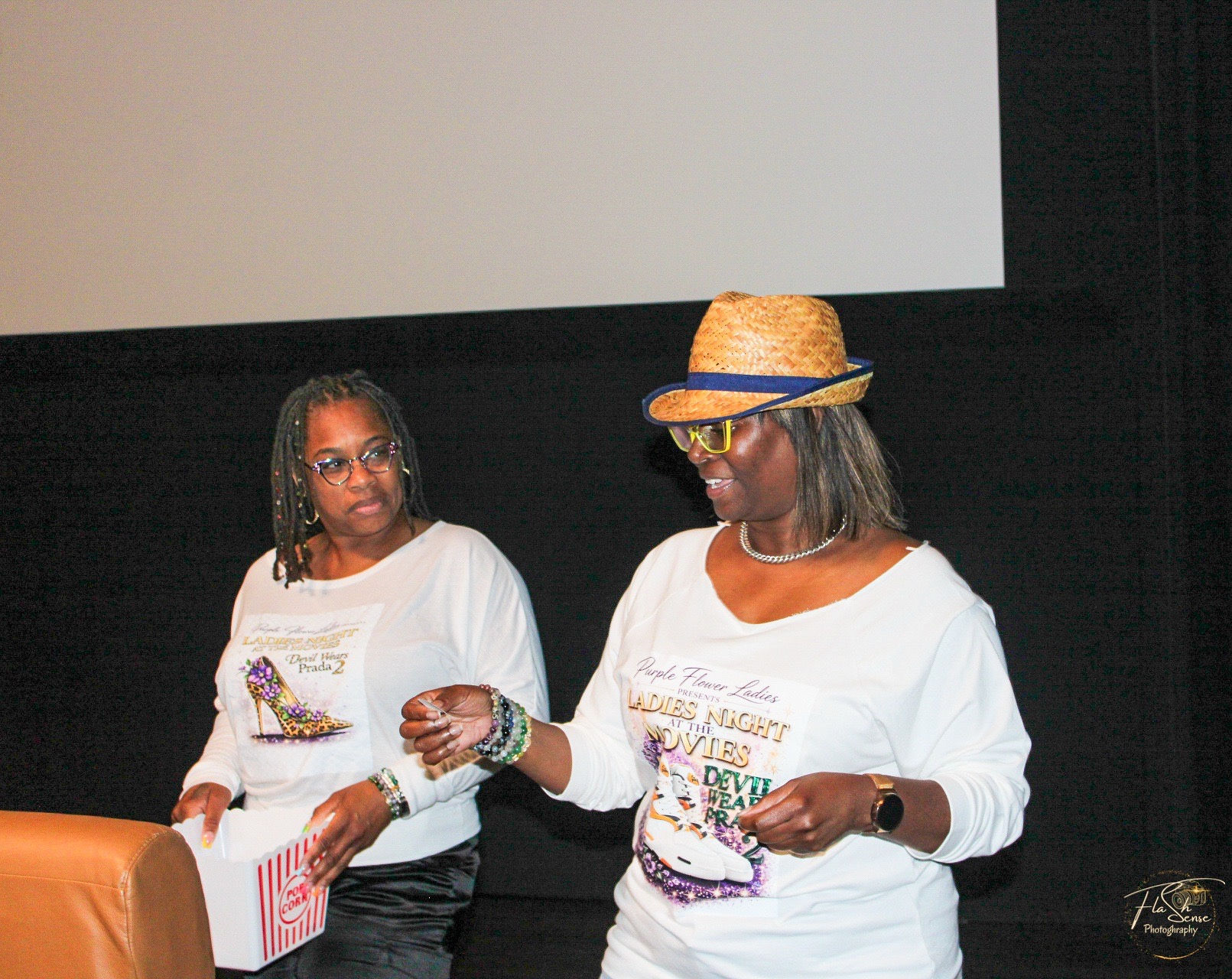 Two women standing on stage, one wearing glasses and a white shirt, the other wearing a straw hat and glasses, holding papers.