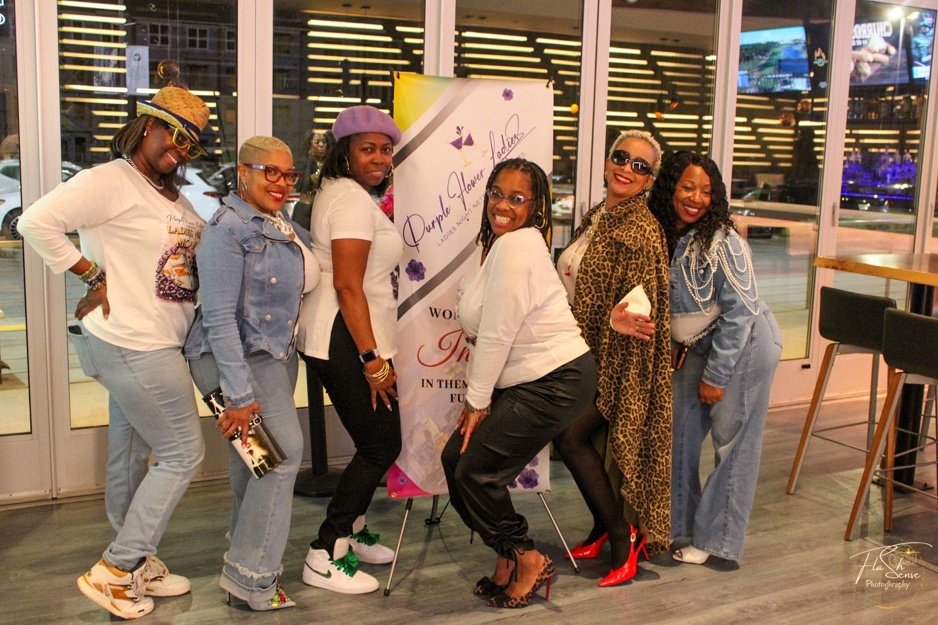 Group of seven women smiling indoors, some wearing glasses and colorful outfits, standing in front of a banner and large windows.