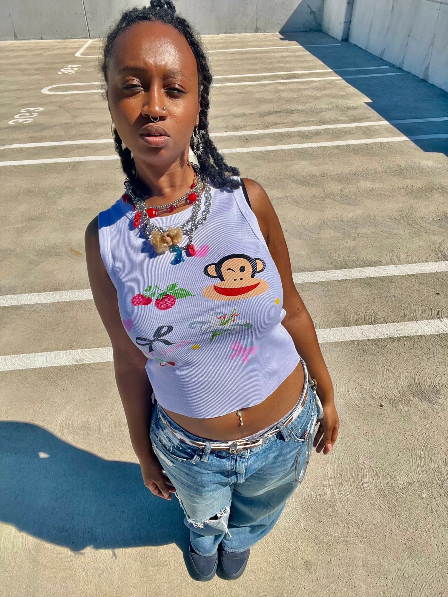 Young woman with braided hair standing outdoors in a parking lot, wearing a white tank top with colorful monkey and strawberry designs, and ripped jeans.