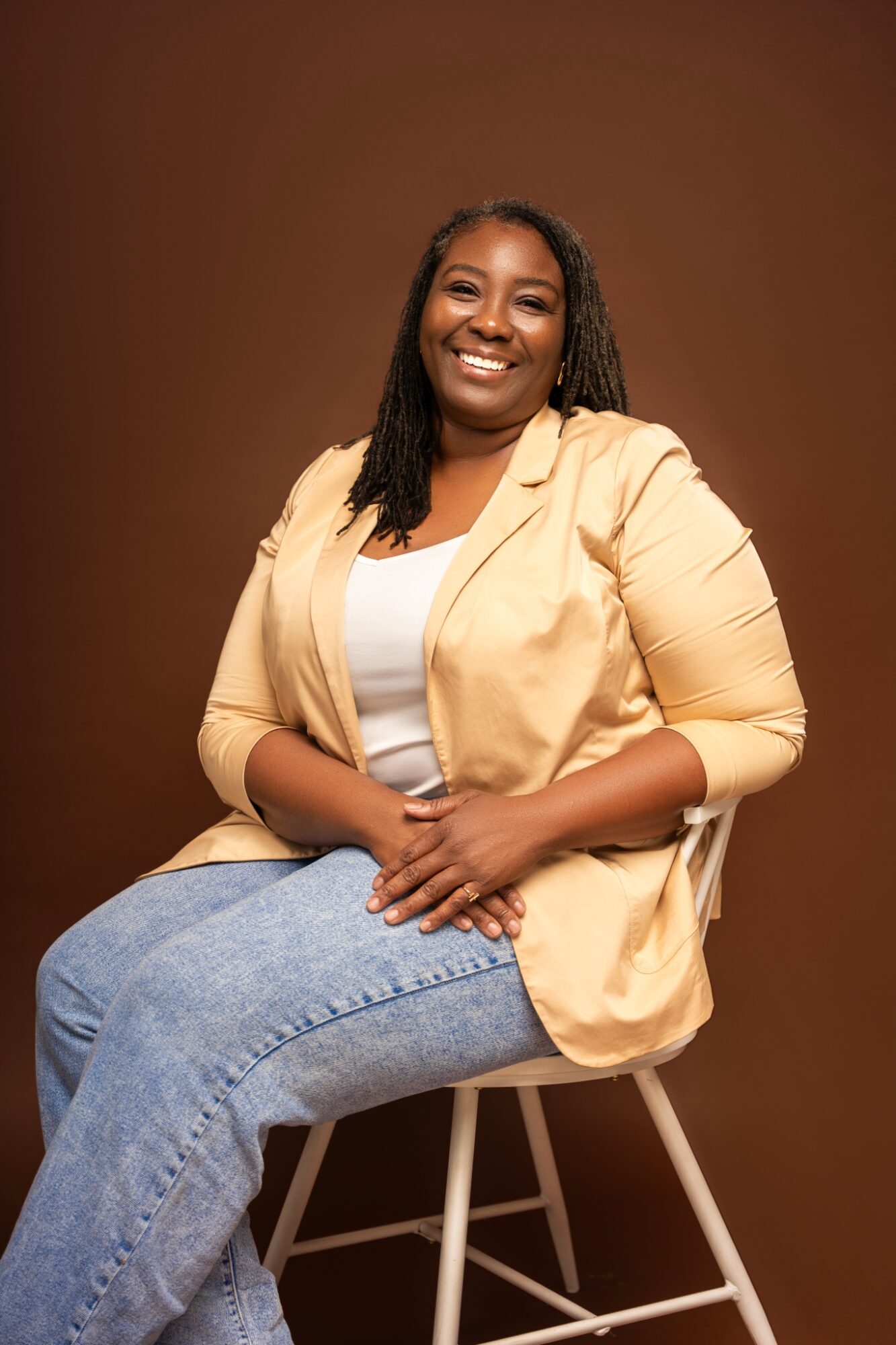 Smiling woman sitting on a stool, wearing a beige blazer and jeans, against a brown background.