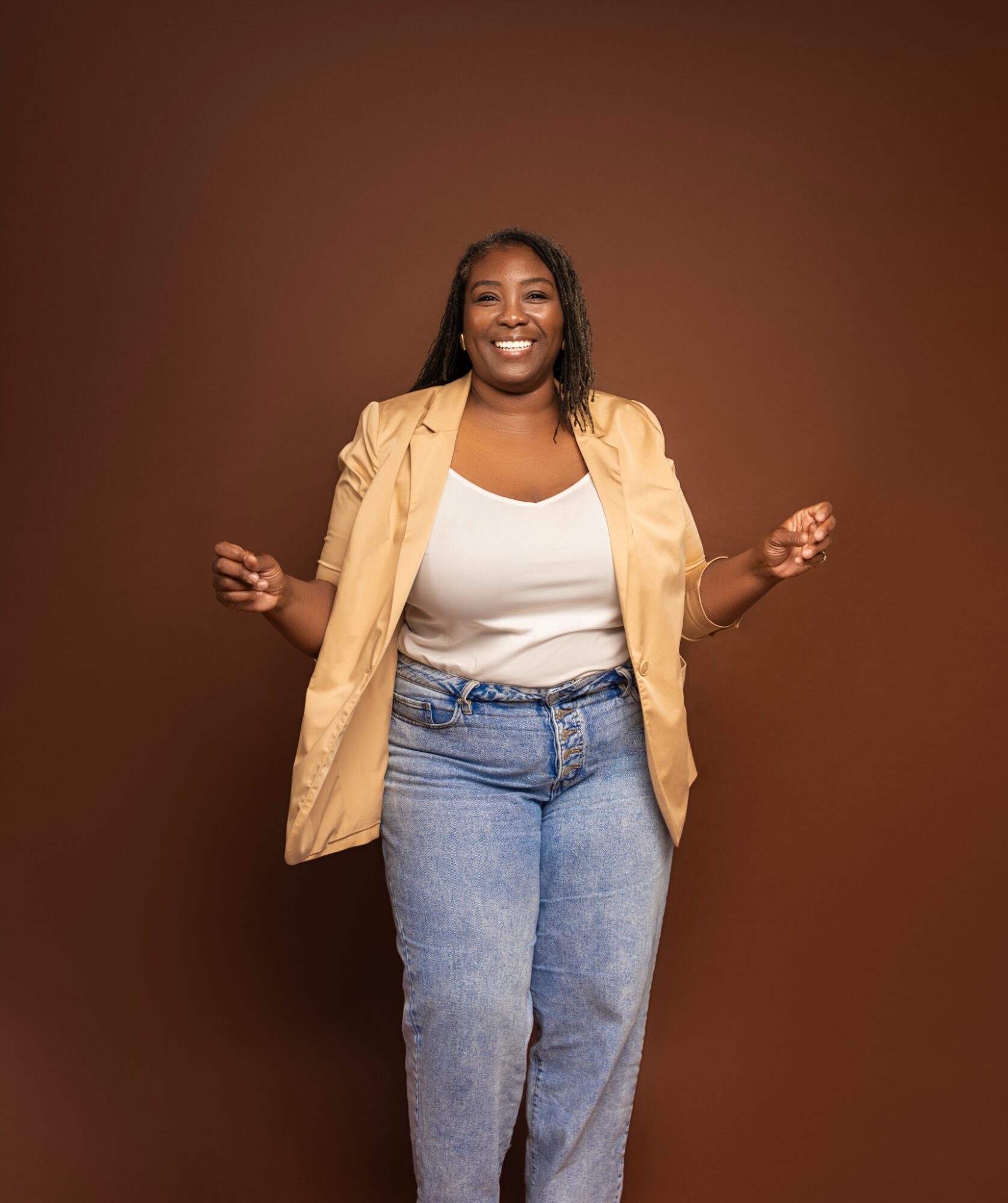 Smiling woman standing with arms slightly raised, wearing a beige blazer, white top, and jeans, against a brown background.