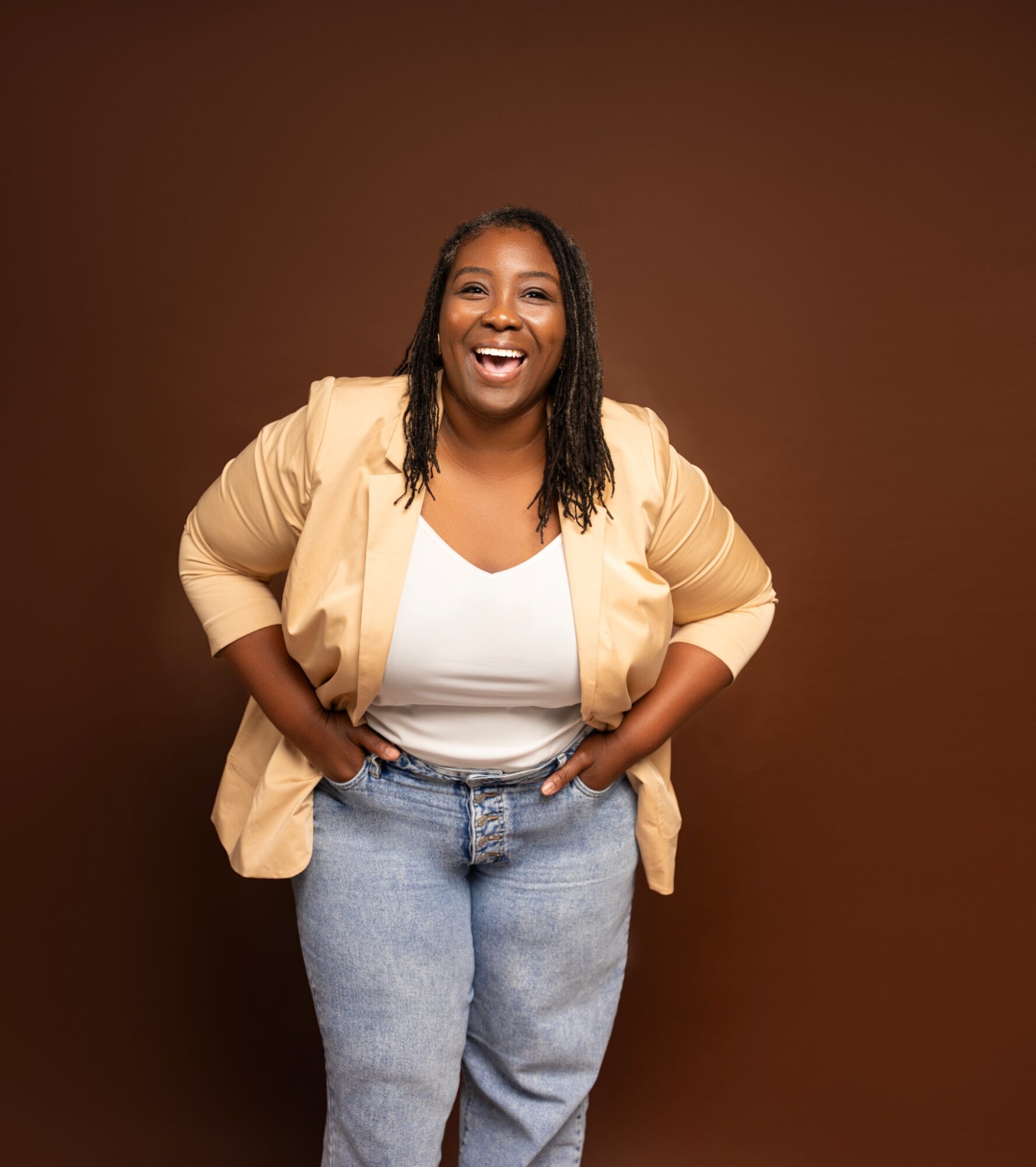 Smiling woman with long hair, wearing a beige blazer and jeans, standing against a brown background.