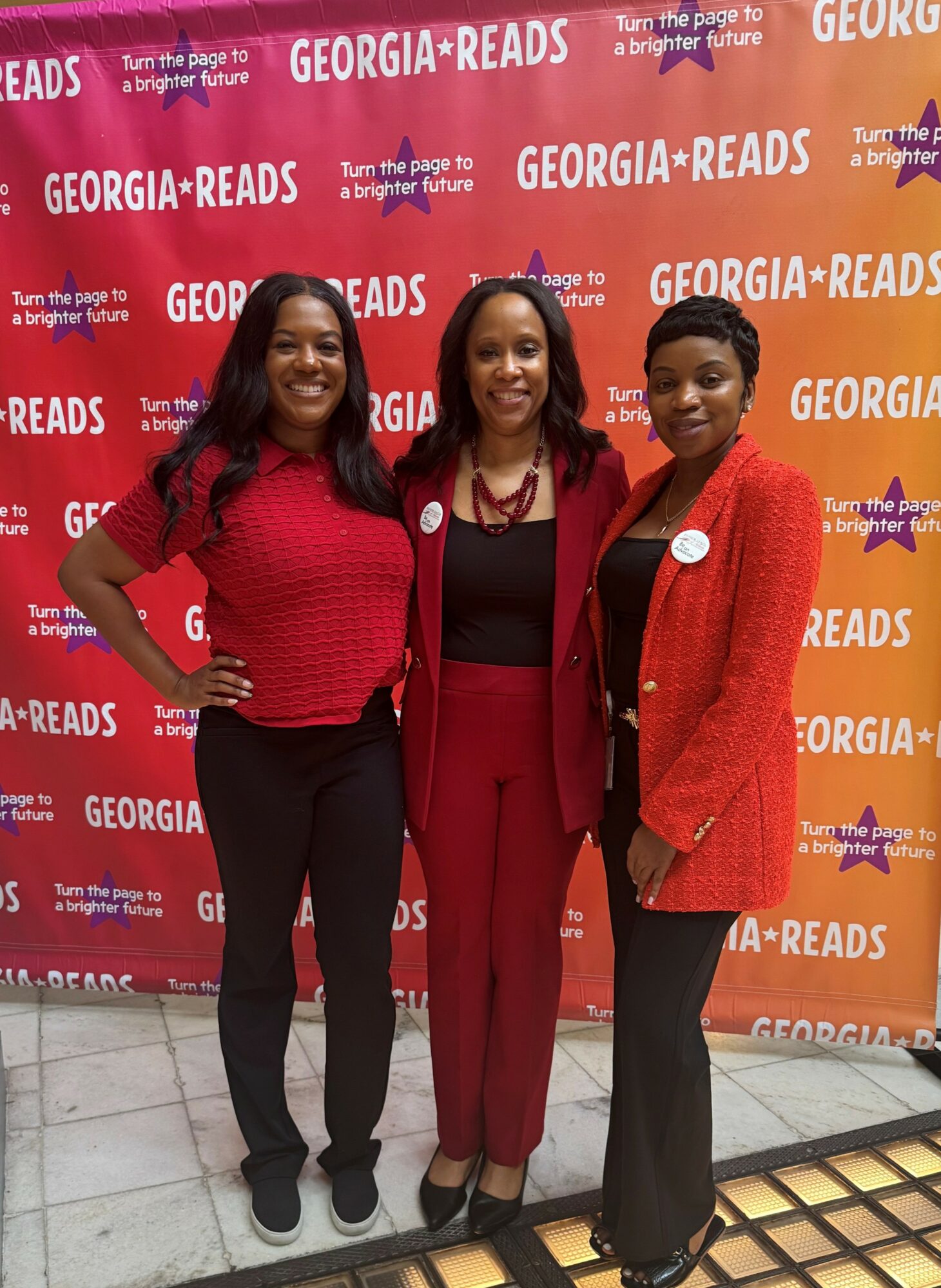 Three women standing in front of a red backdrop with white text, smiling, dressed in red and black clothing.