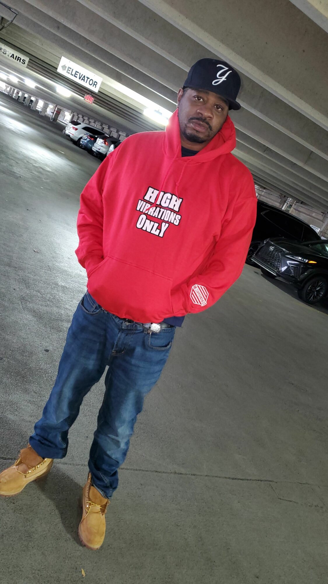 Person standing in parking garage wearing a red hoodie, jeans, tan boots, and a black cap.