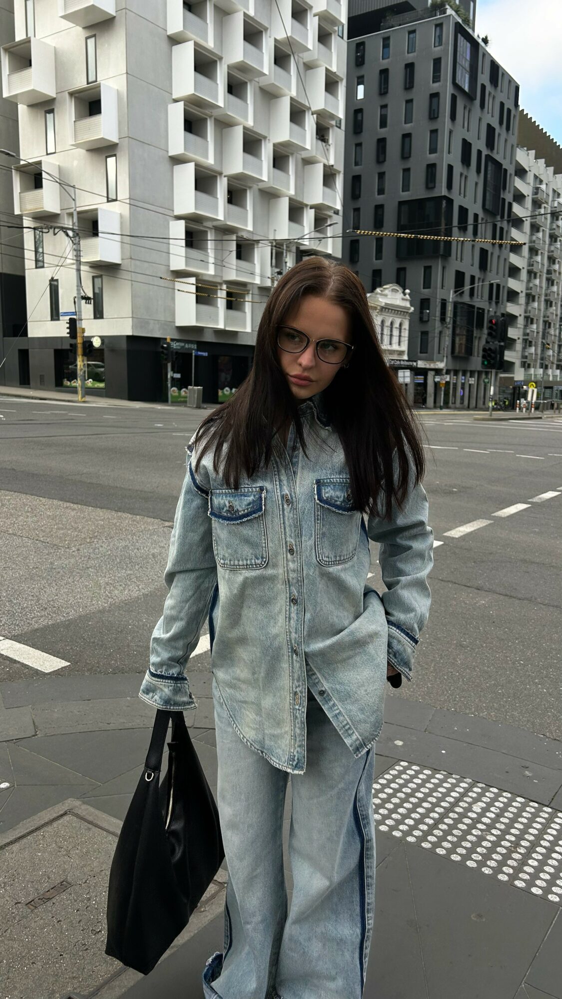 Young woman with long dark hair in a denim jacket and pants holding a black bag stands on city street.