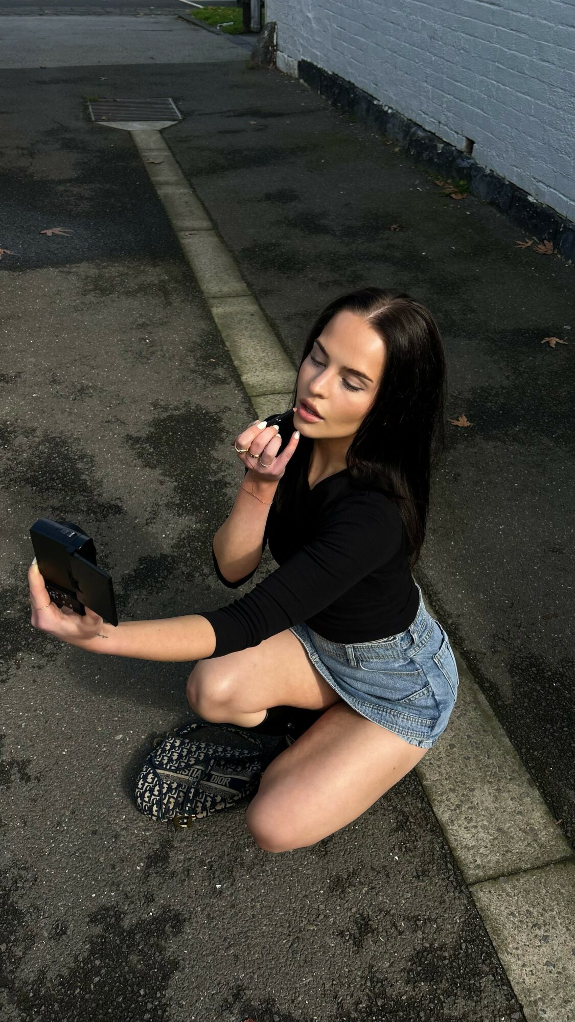 Young woman with long dark hair kneeling on pavement, holding a phone, wearing black top and denim shorts.
