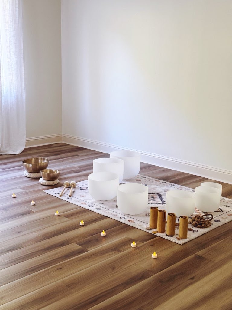 Decorative setup with candles, bowls, and small objects on a rug on wooden floor in a room with white walls.