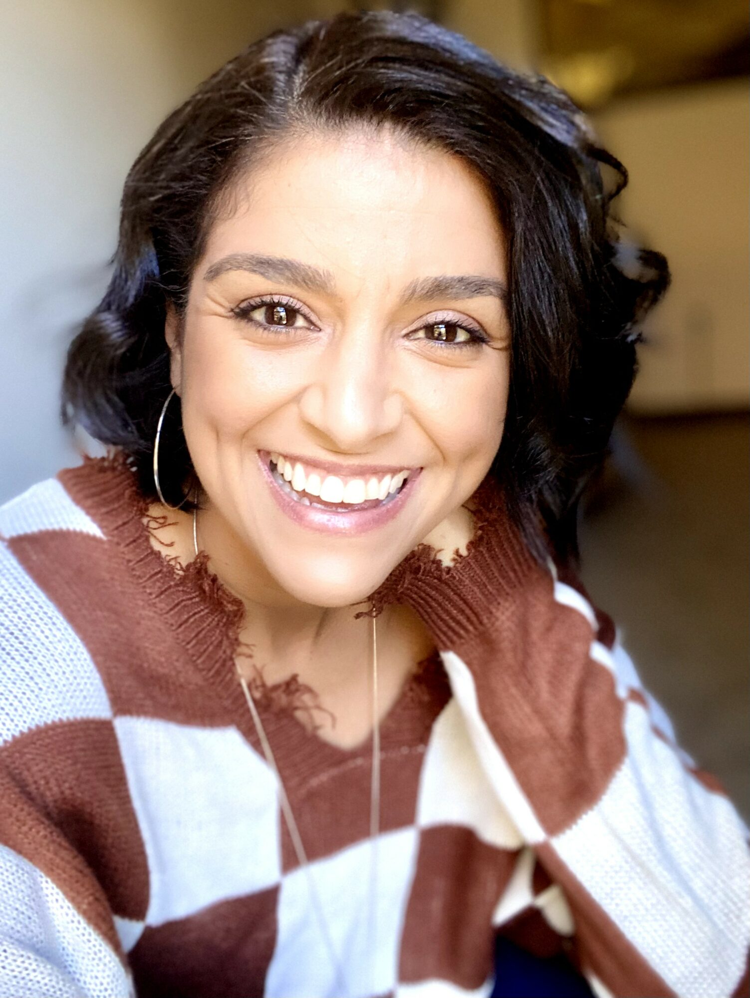 Smiling woman with dark wavy hair, wearing a checkered sweater, in a well-lit indoor setting.