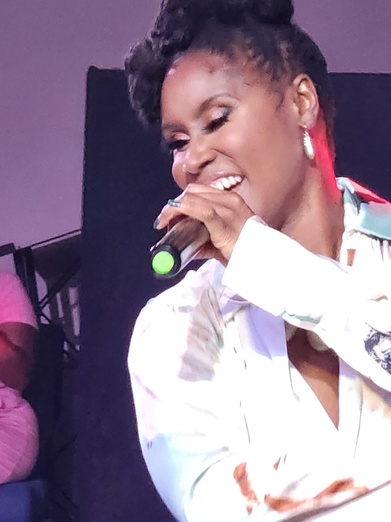 Woman singing into a microphone, smiling, wearing a white shirt and red earrings, with a dark background.