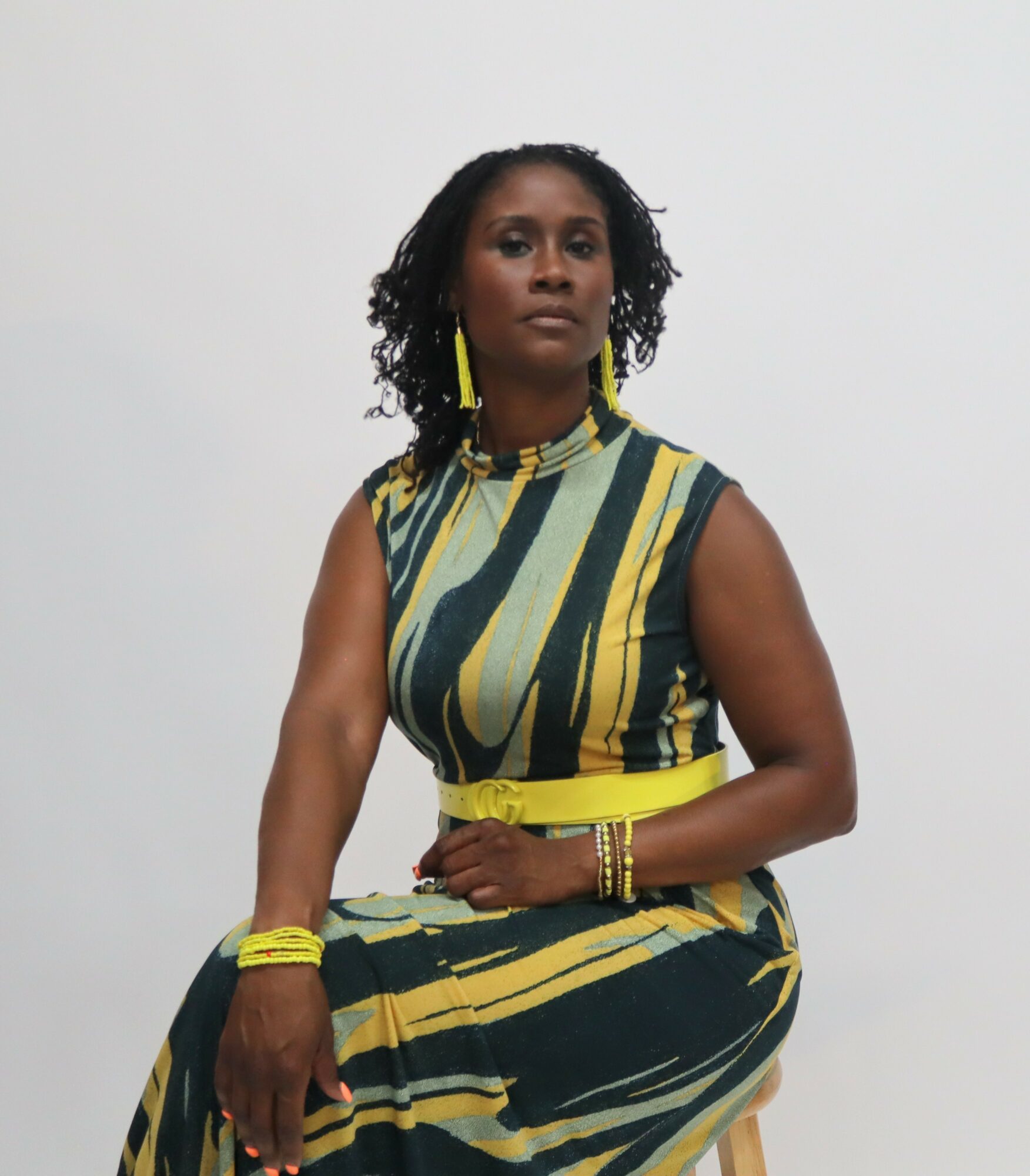 Woman with curly hair in a sleeveless striped dress, sitting against a plain background.