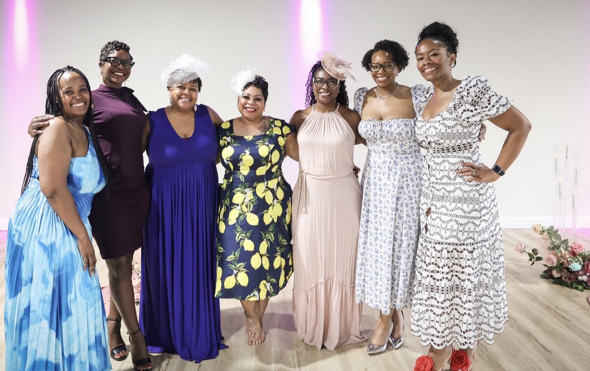 Seven women standing together indoors, smiling, dressed in colorful dresses, with a plain background and pink lighting.