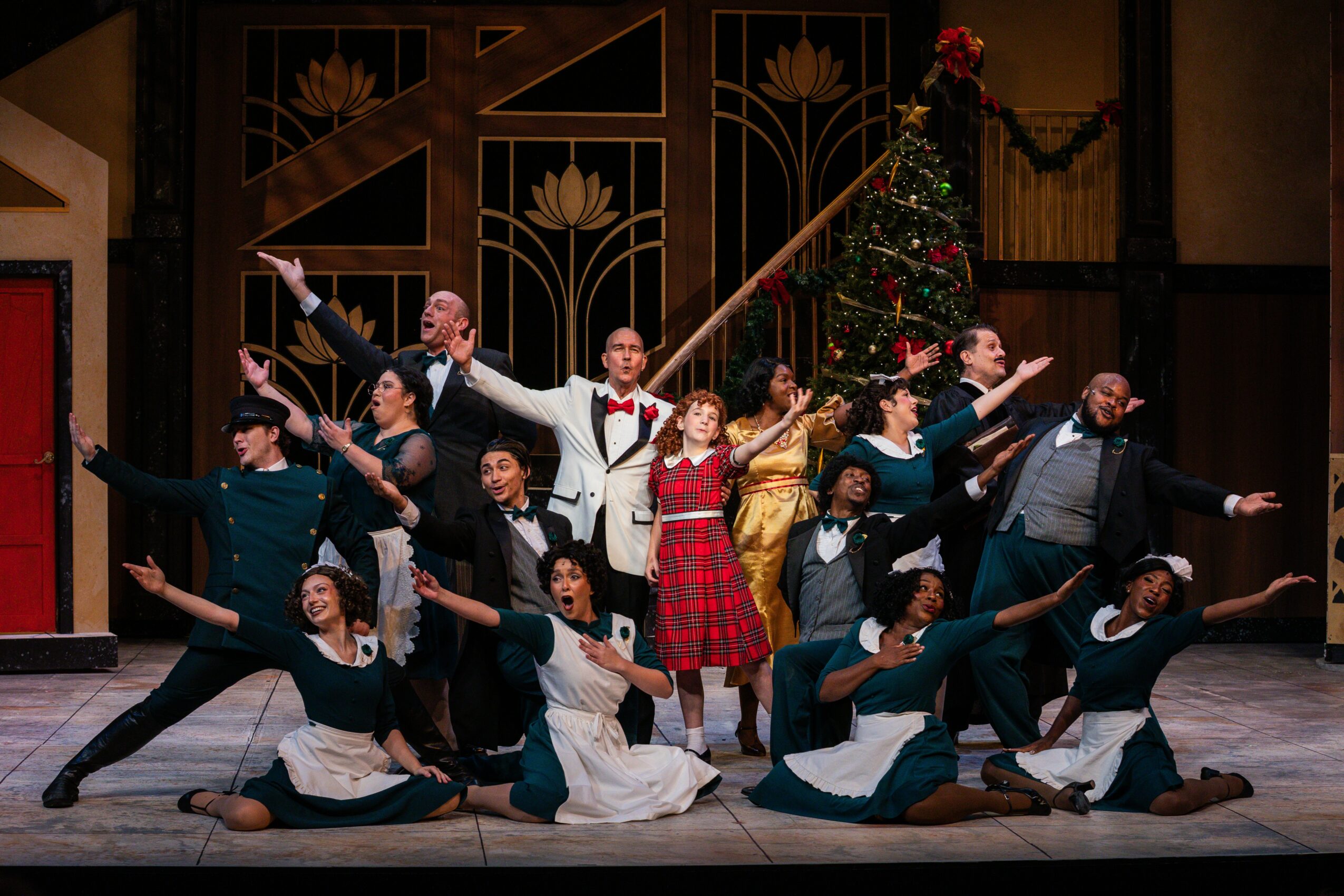 Group of performers on stage, some standing and some kneeling, with a Christmas tree and decorated backdrop behind them.