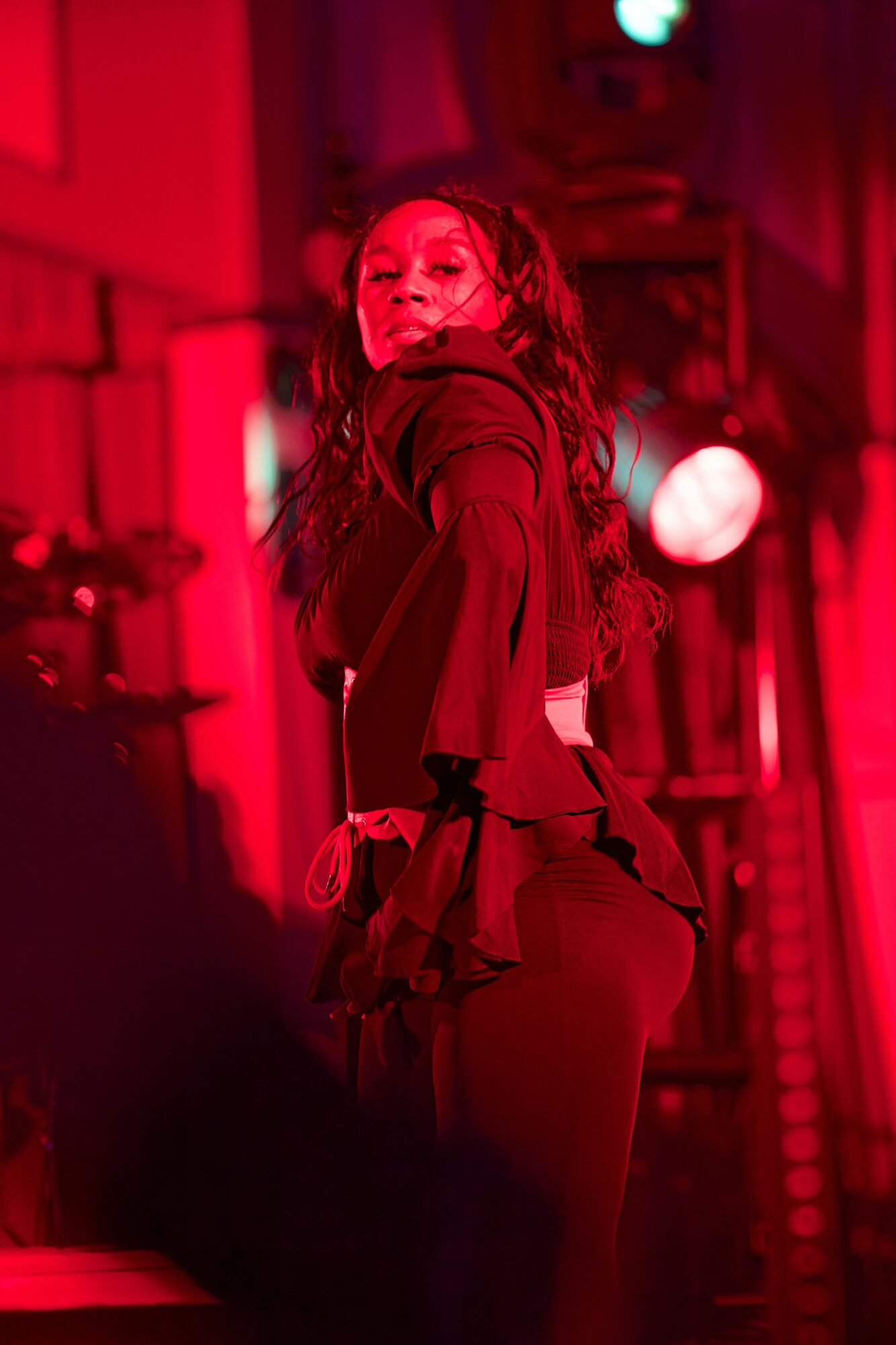 A woman with long hair in a dark outfit stands on stage with red lighting, looking over her shoulder.