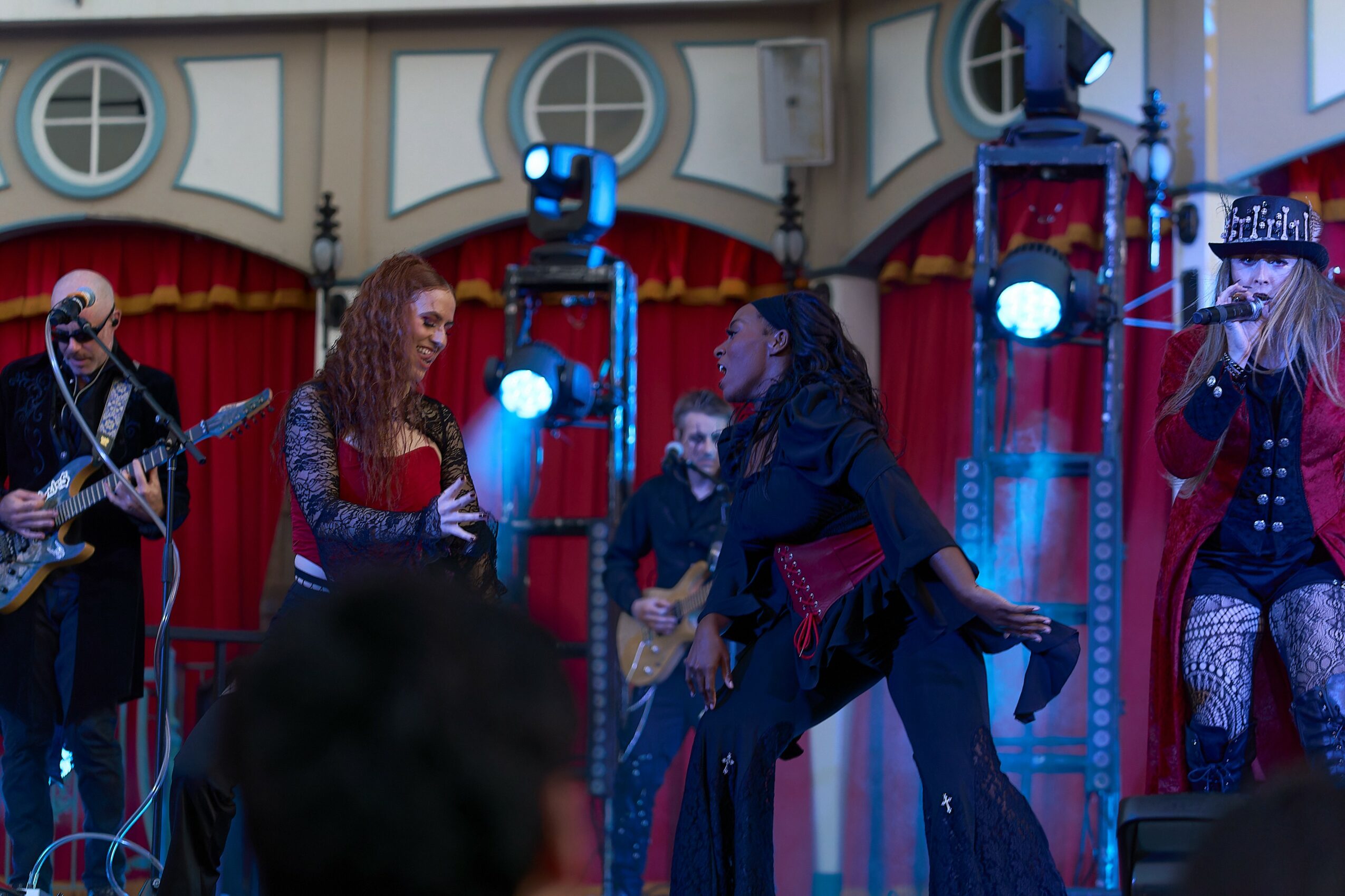 Group of musicians performing on stage with red curtains and blue lighting, including a singer and guitarists.