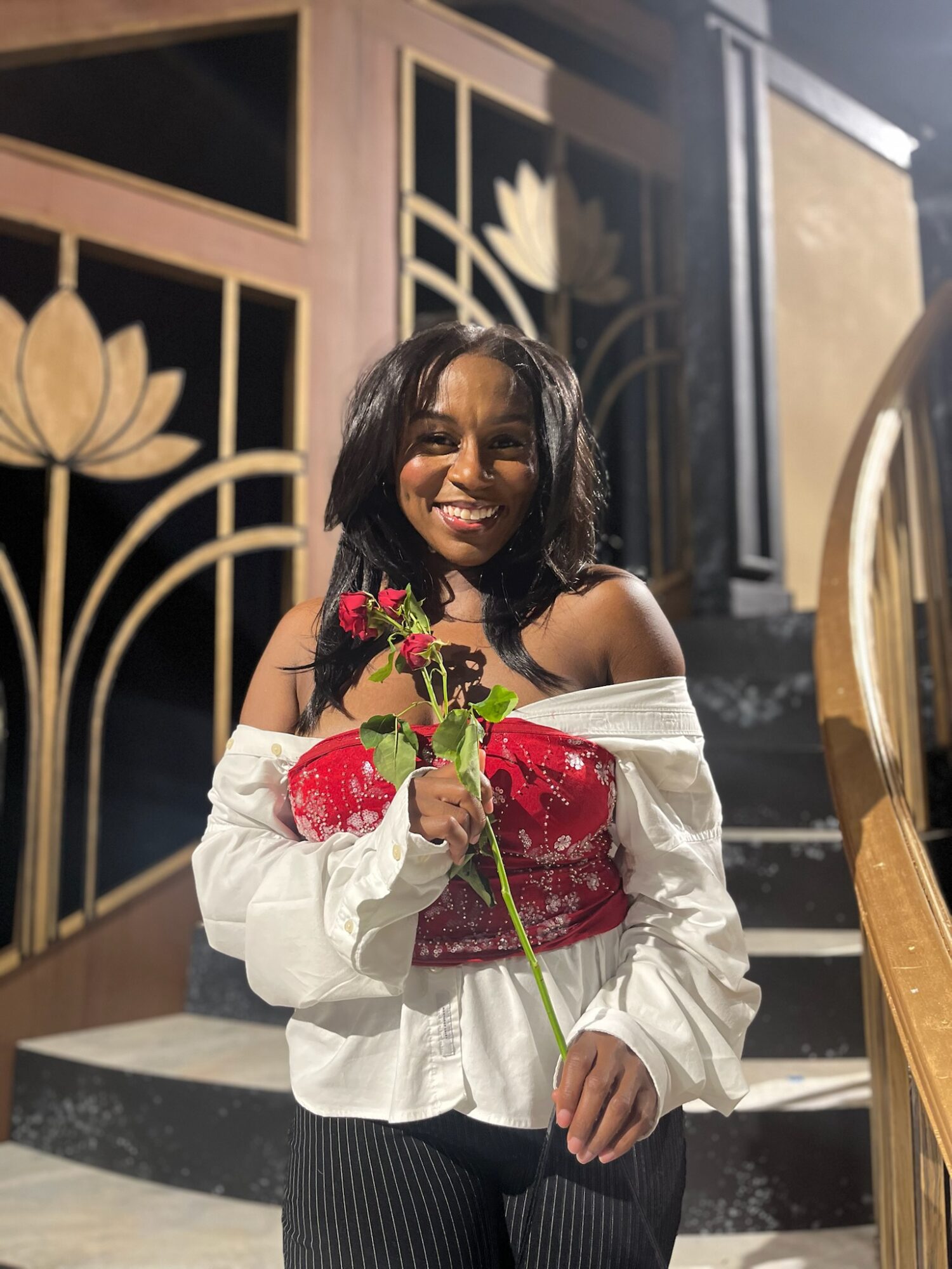Smiling woman holding a flower, standing indoors near staircase and decorative wooden panels.