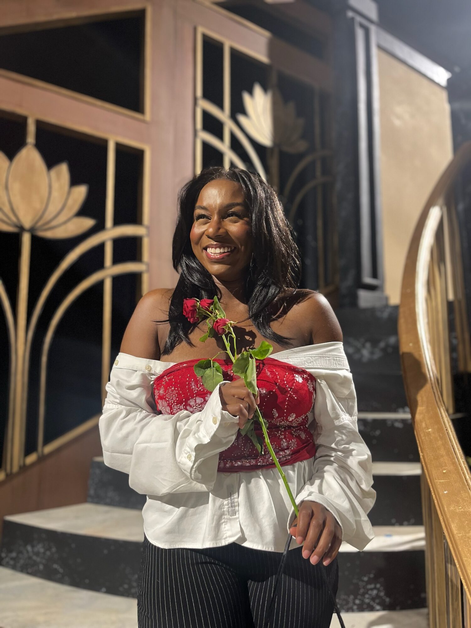 Woman smiling, holding a red rose, standing indoors with decorative gold and black background.