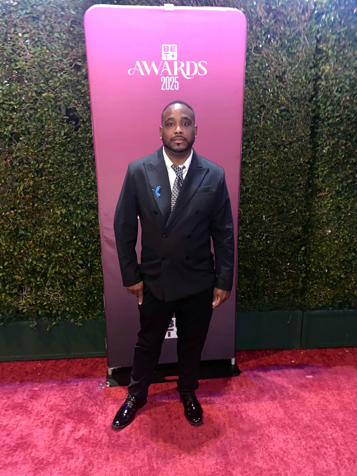 Man in a dark suit and tie standing on red carpet in front of pink backdrop with awards logo, facing forward.