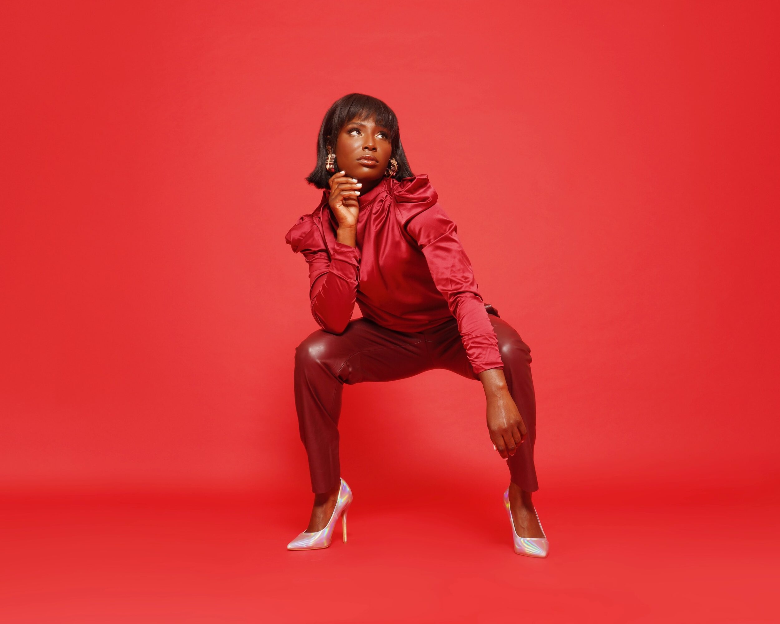 Woman in red outfit squatting with hand near chin against red background.