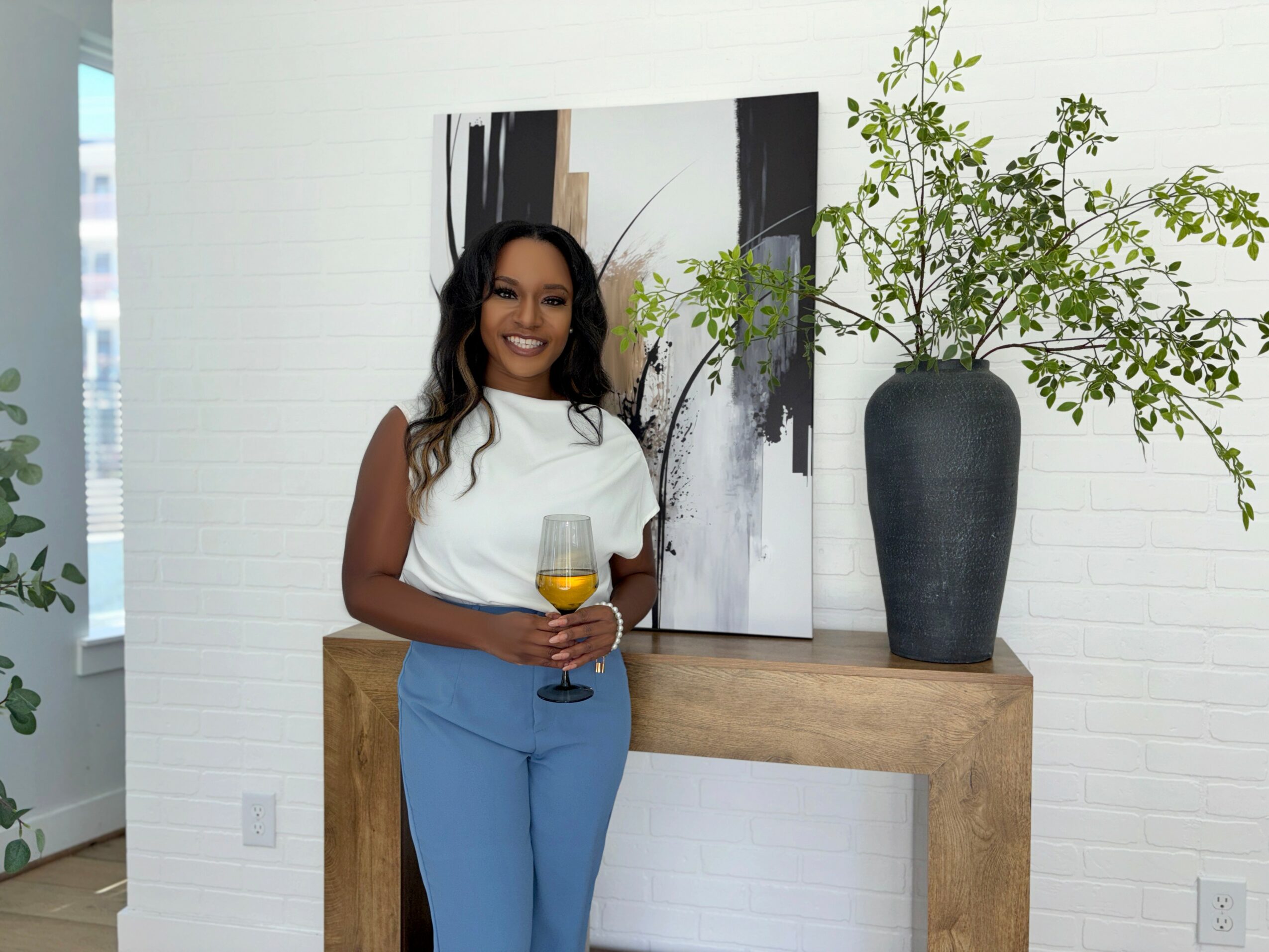 Woman with long dark hair holding a glass of wine, standing next to a large vase with green plant, in a bright room.