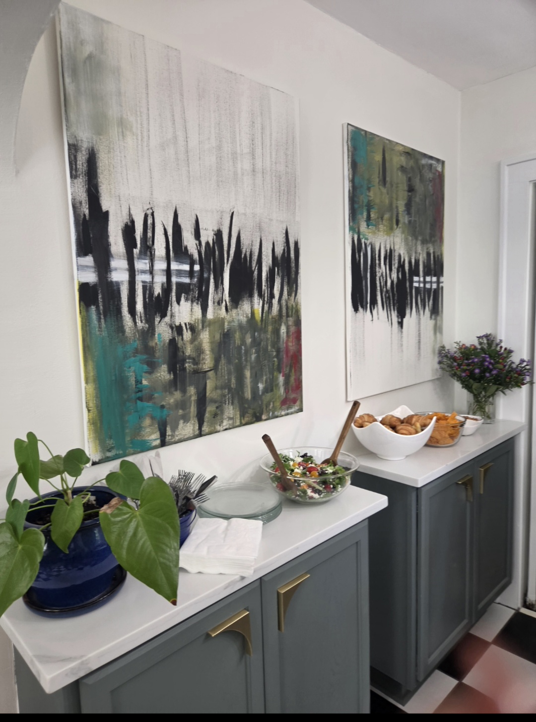 Two abstract paintings hang above a blue cabinet with decorative items, including a plant, bowl, and flowers, in a bright room.
