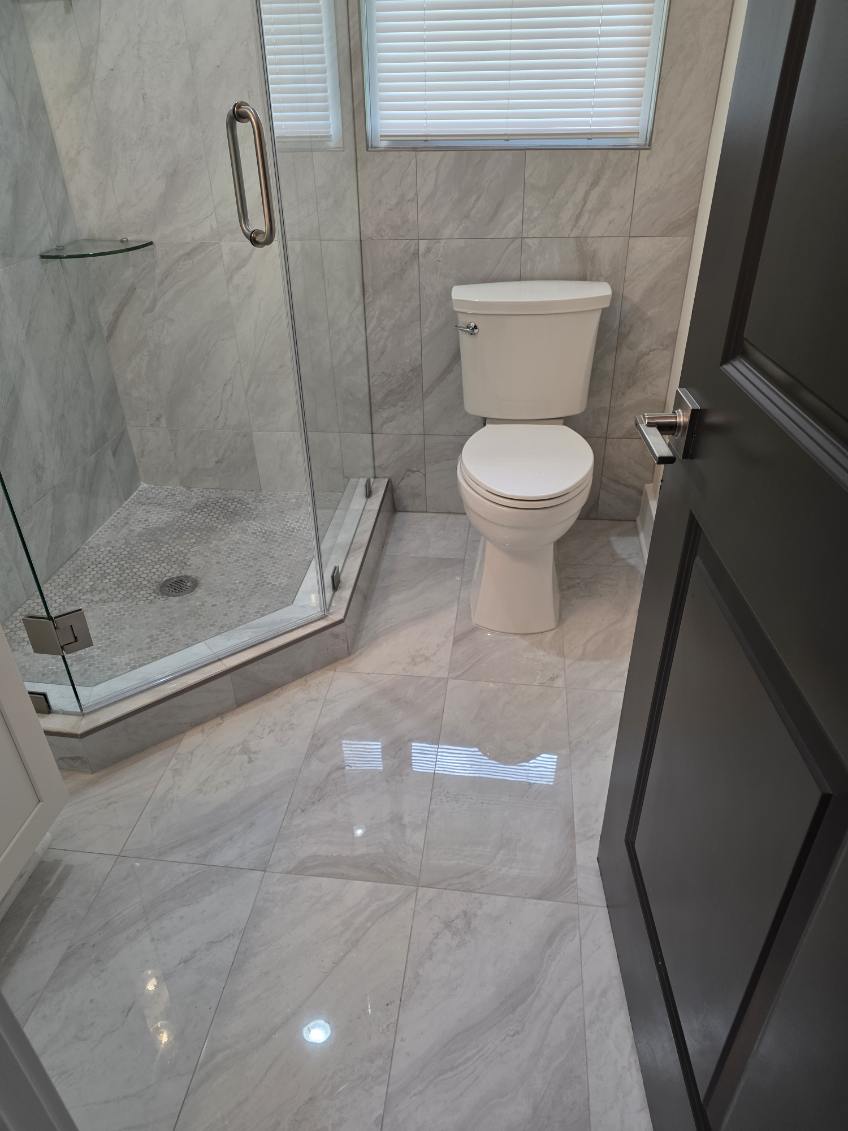 Bathroom with shower enclosure and toilet, marble floor and walls, window with blinds, door partially open.