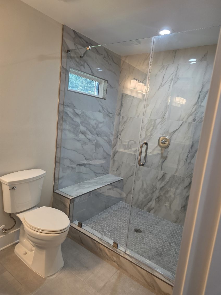 Bathroom with a toilet and a glass-enclosed shower with marble walls and a small window.