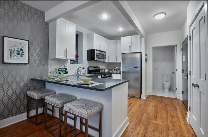 Kitchen with white cabinets, stainless steel appliances, a small island with three stools, and a view of a bathroom in the background.