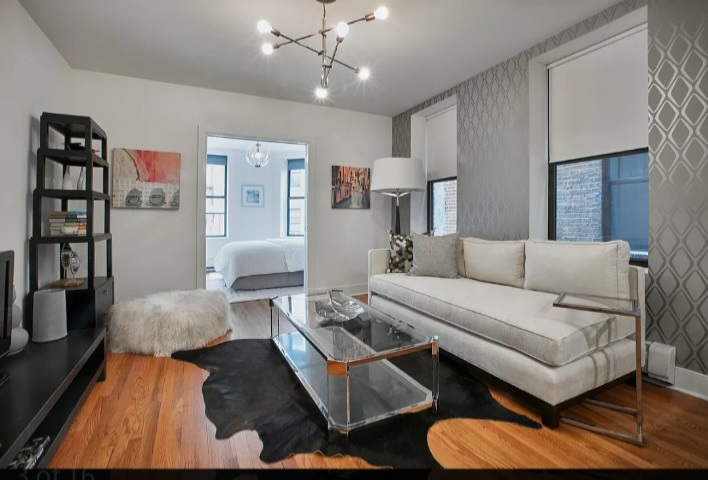 Living room with white sofa, black bookshelf, glass coffee table, and a cowhide rug, with a bedroom visible through an open doorway.