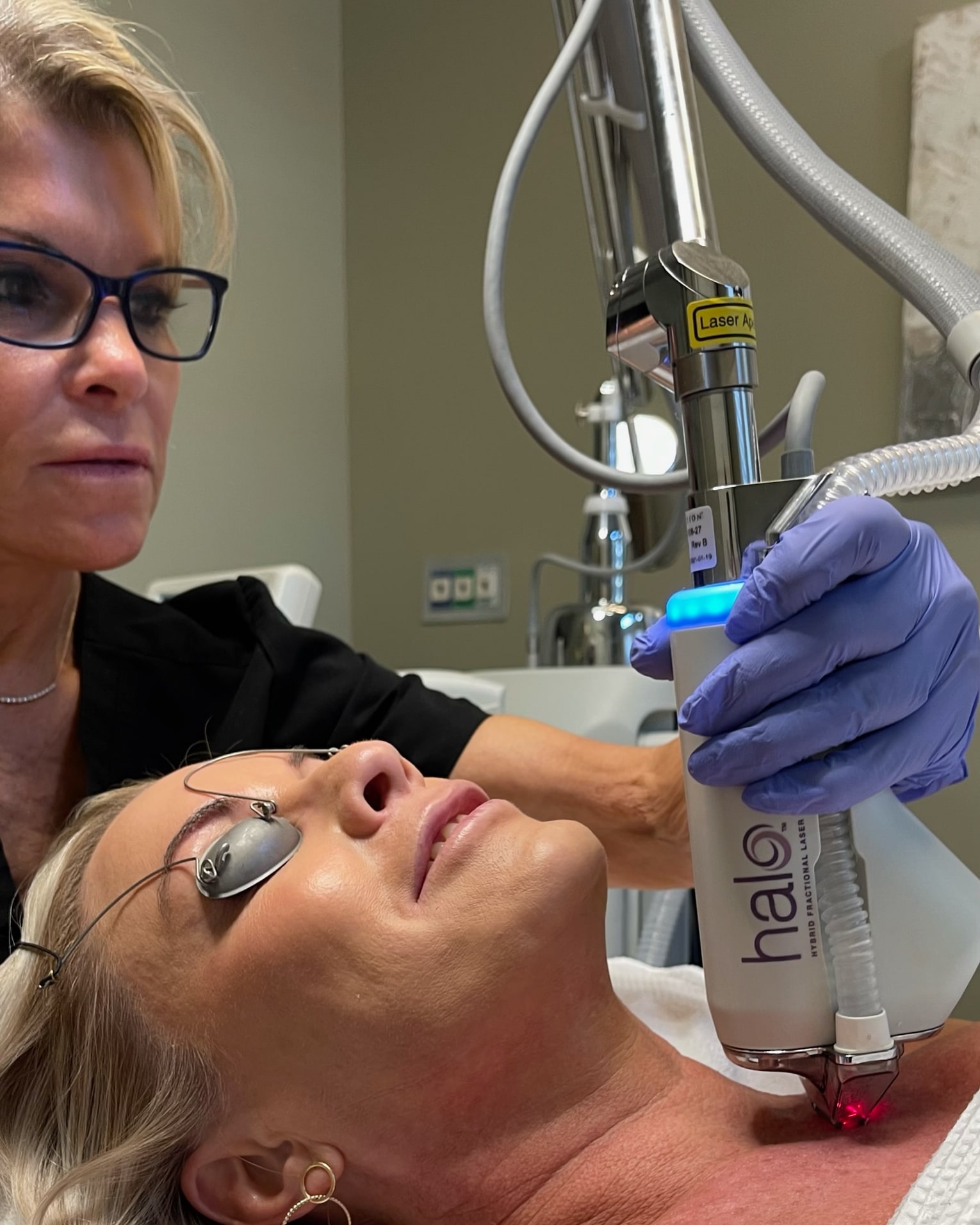 Medical professional in gloves performs a laser treatment on a woman lying down with eye protection, in a clinical setting.