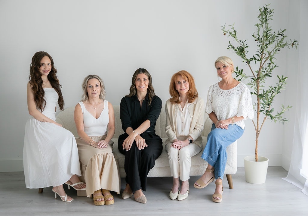 Five women sitting on a white bench in a bright room with a potted plant, dressed in light-colored clothing.