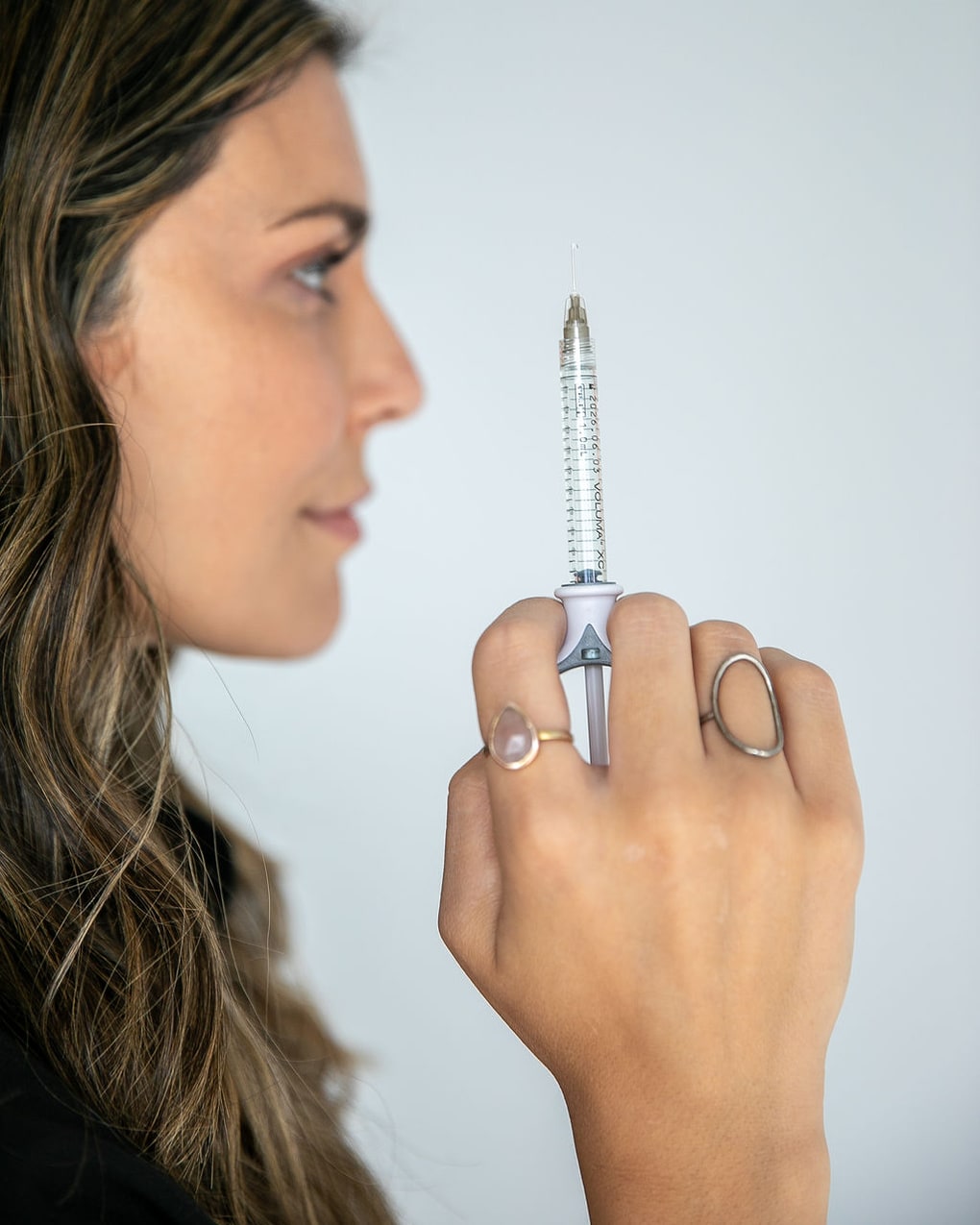 Side profile of a woman holding a syringe, with focus on her face and hand, against a plain background.