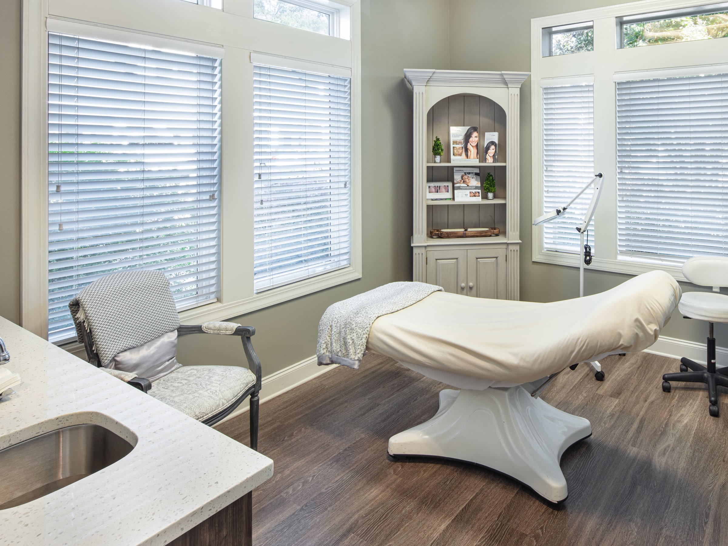 Medical examination room with a bed, chair, cabinet, and large windows with blinds, bright and clean space.