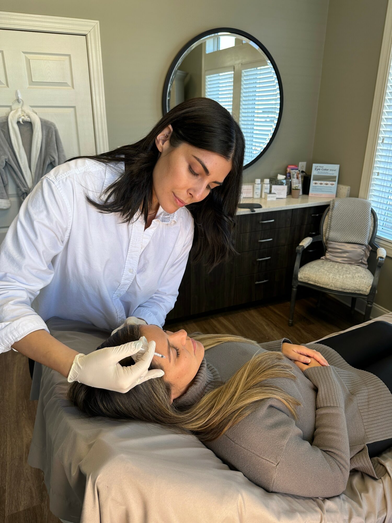 A woman in a white shirt performs a facial treatment on a woman lying on a bed in a treatment room.