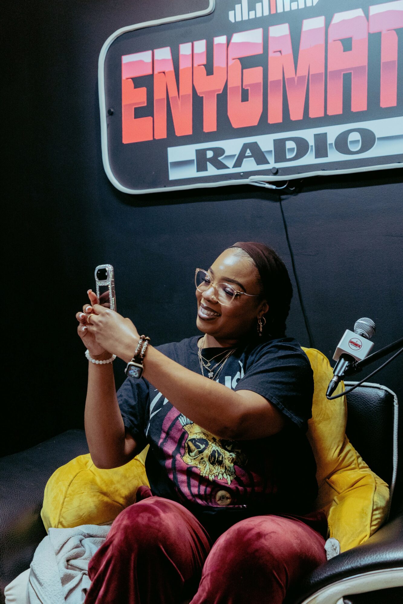 Woman sitting on yellow cushion, smiling, taking a selfie with a phone, in front of a radio station sign, wearing glasses and a dark shirt.